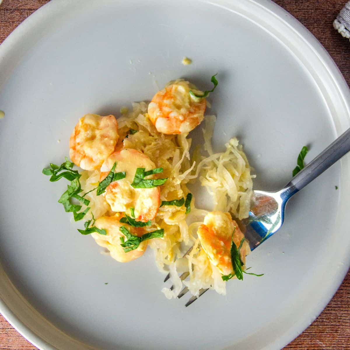 A fork holds Dijon-sauced shrimp with herbs over sauerkraut on a light gray plate.