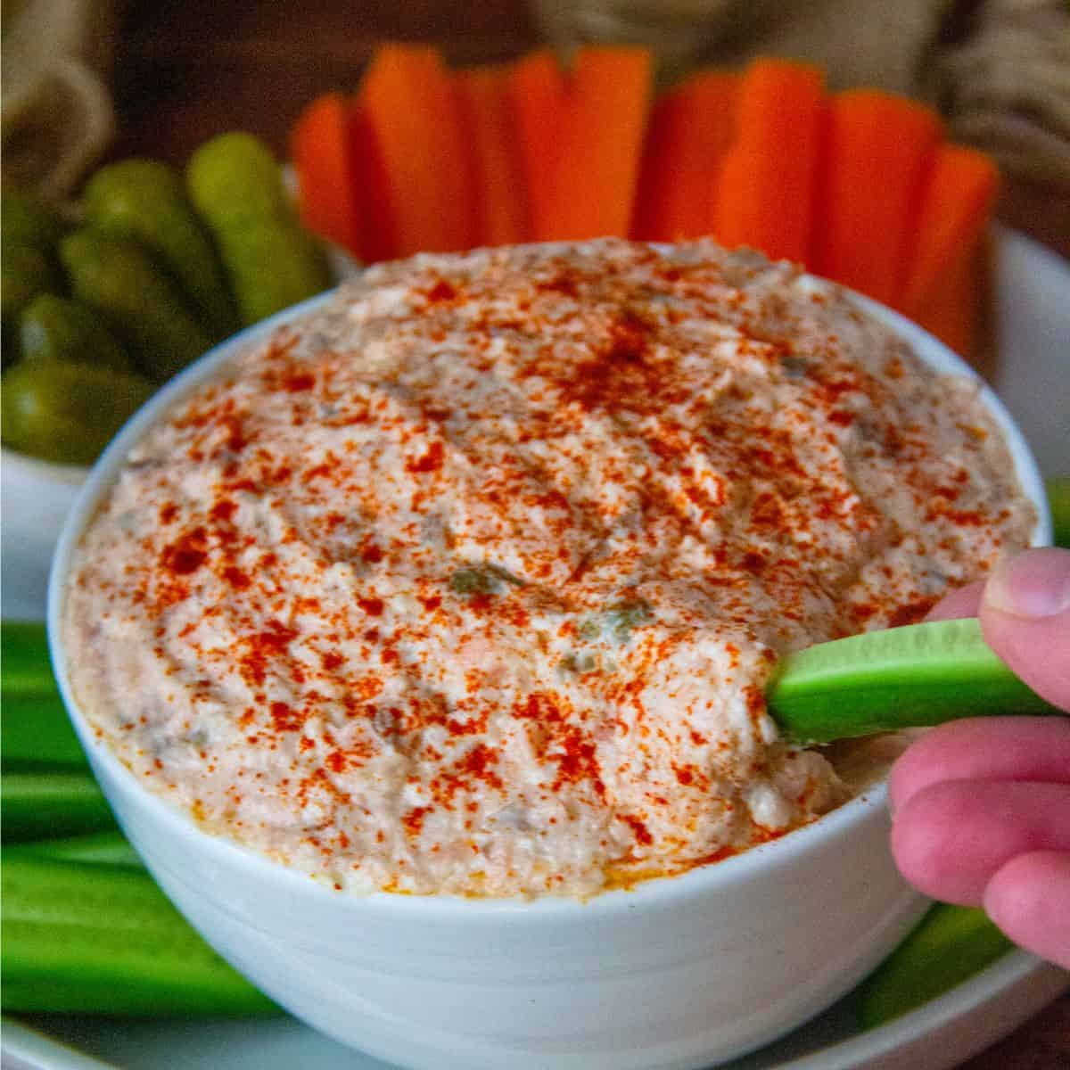 Hand dips a celery stick into a bowl of salmon spread topped with paprika, ringed by carrots.
