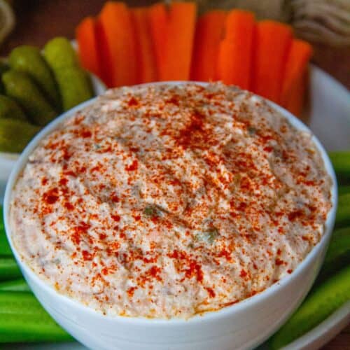 Close-up of paprika-dusted salmon spread in a white bowl, surrounded by pickles and carrots.