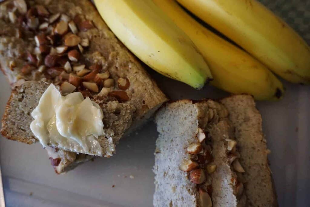 Keto banana bread with buttered slice on cutting board next to whole bananas.