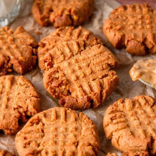 Peanut butter cookies with crisscross fork marks on parchment paper, beside a spoonful of peanut butter.