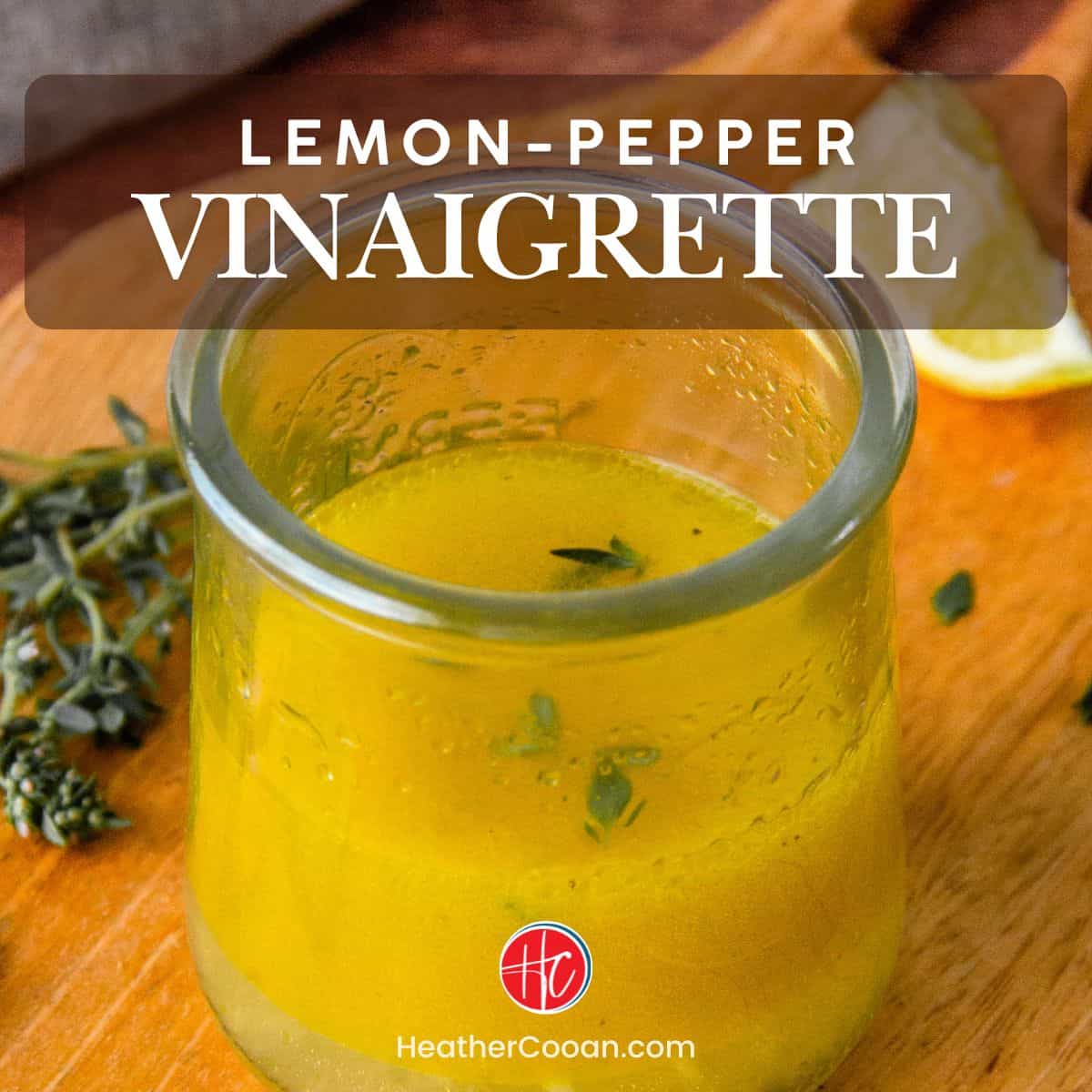 Lemon pepper vinaigrette in a glass jar with fresh thyme and lemon wedges on a wood table.