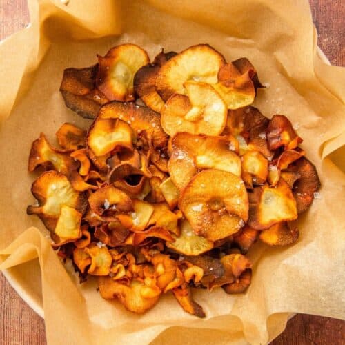 A parchment-lined bowl holds golden and dark brown parsnip chips sprinkled with coarse salt.
