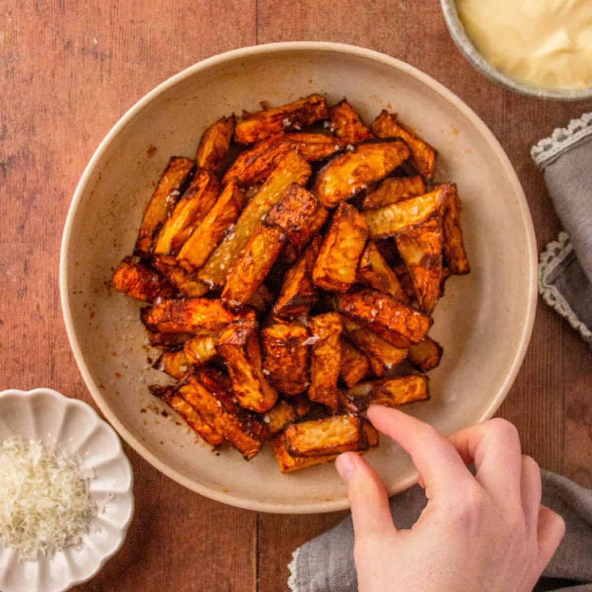 A hand reaches for golden, dark-edged celery root fries in a bowl, cheese and dip nearby.