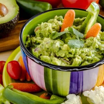 Fresh chunky guacamole in colorful bowl sitting on bright green plate garnished with raw carrots and green peppers and cilantro surrounded by raw vegetables