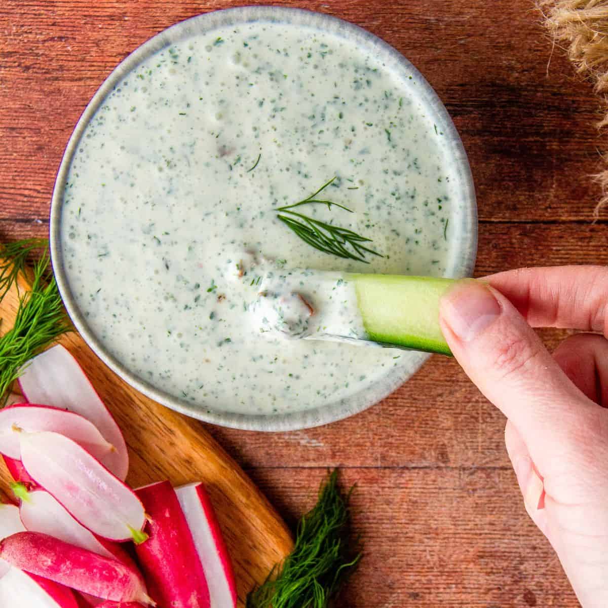 A hand dips a cucumber stick into creamy dill dressing beside sliced radishes and fresh dill.
