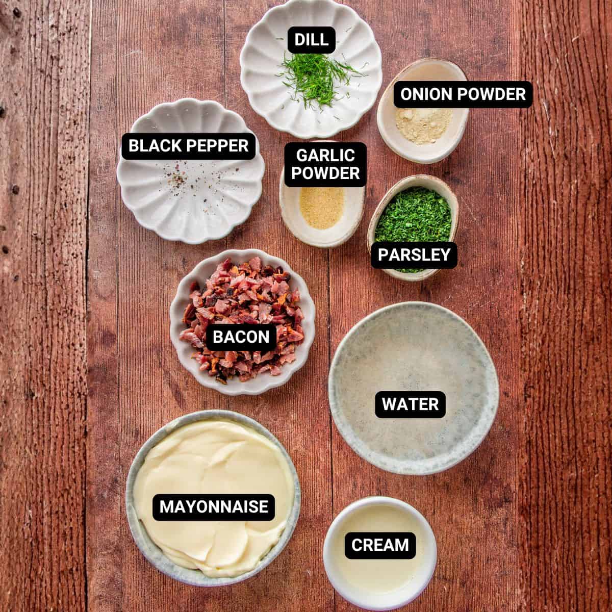 Labeled bowls of dill, bacon, mayonnaise, cream, parsley, and spices on a wood table.