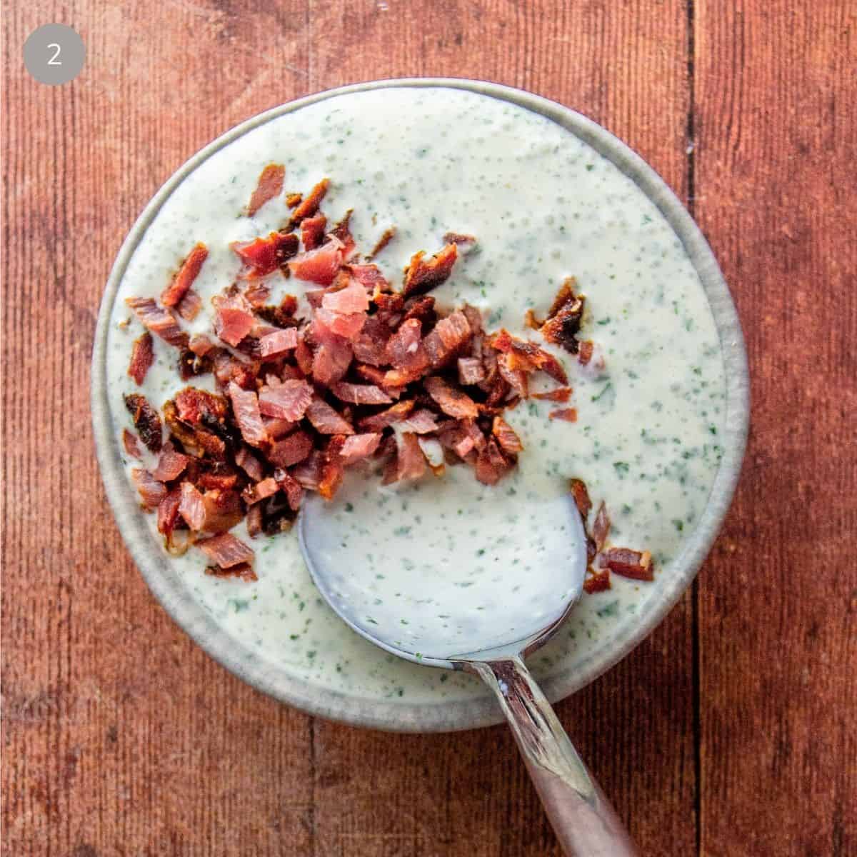 Crispy bacon bits piled on top of smooth green herb dressing in a speckled bowl, with a spoon.