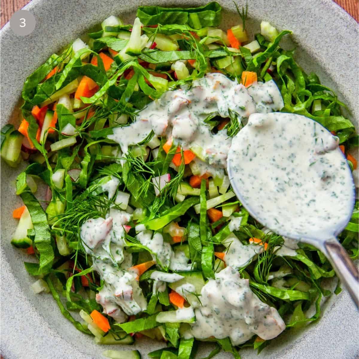 Creamy bacon ranch dressing spooned over a shredded green salad with carrots and radish in a bowl.