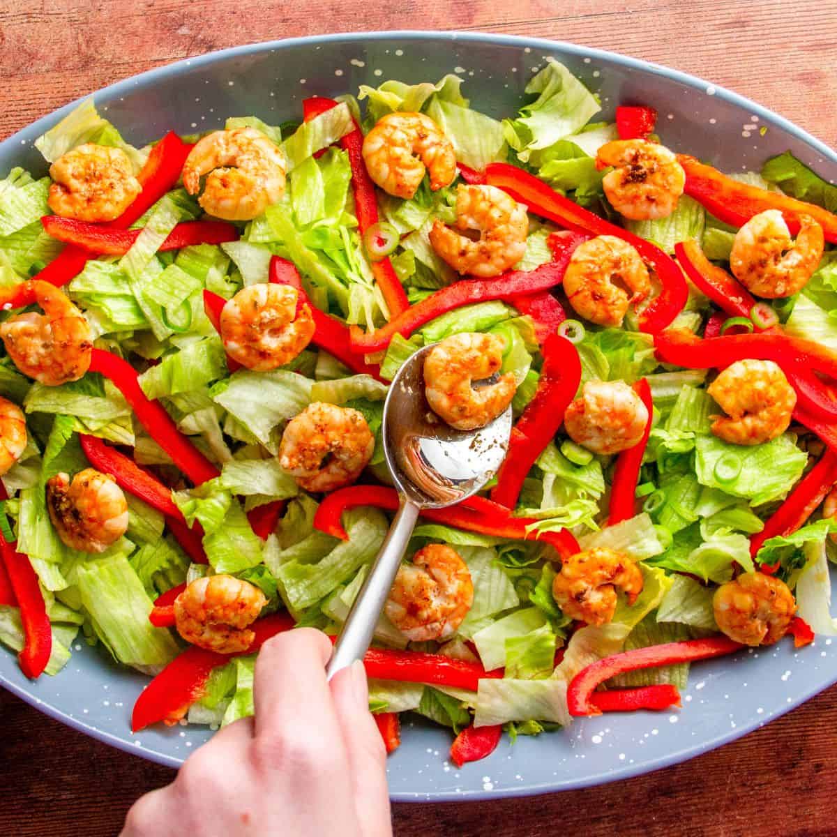 Hand spoons a shrimp onto a platter of lettuce, red pepper, and cilantro salad.