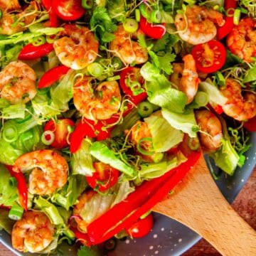 Close-up of shrimp salad with lettuce, red pepper, tomatoes, and sprouts, wooden spoon resting on top.