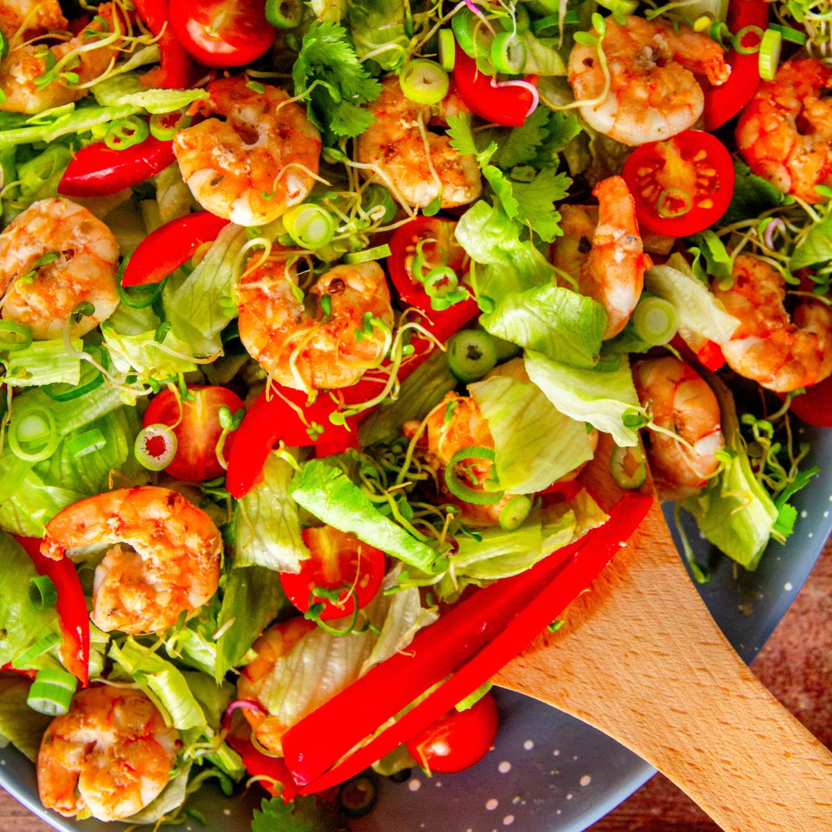 Close-up of shrimp salad with lettuce, red pepper, tomatoes, and sprouts, wooden spoon resting on top.