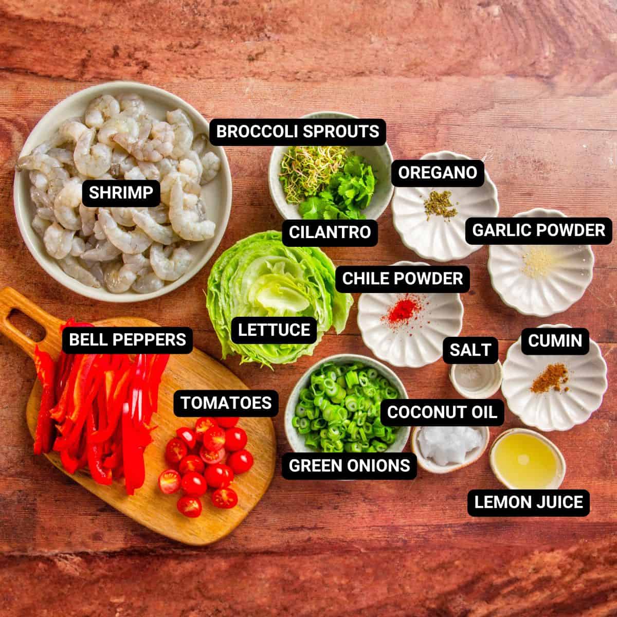 Labeled bowls of shrimp, bell peppers, tomatoes, lettuce, cilantro, and spices on a wood table.