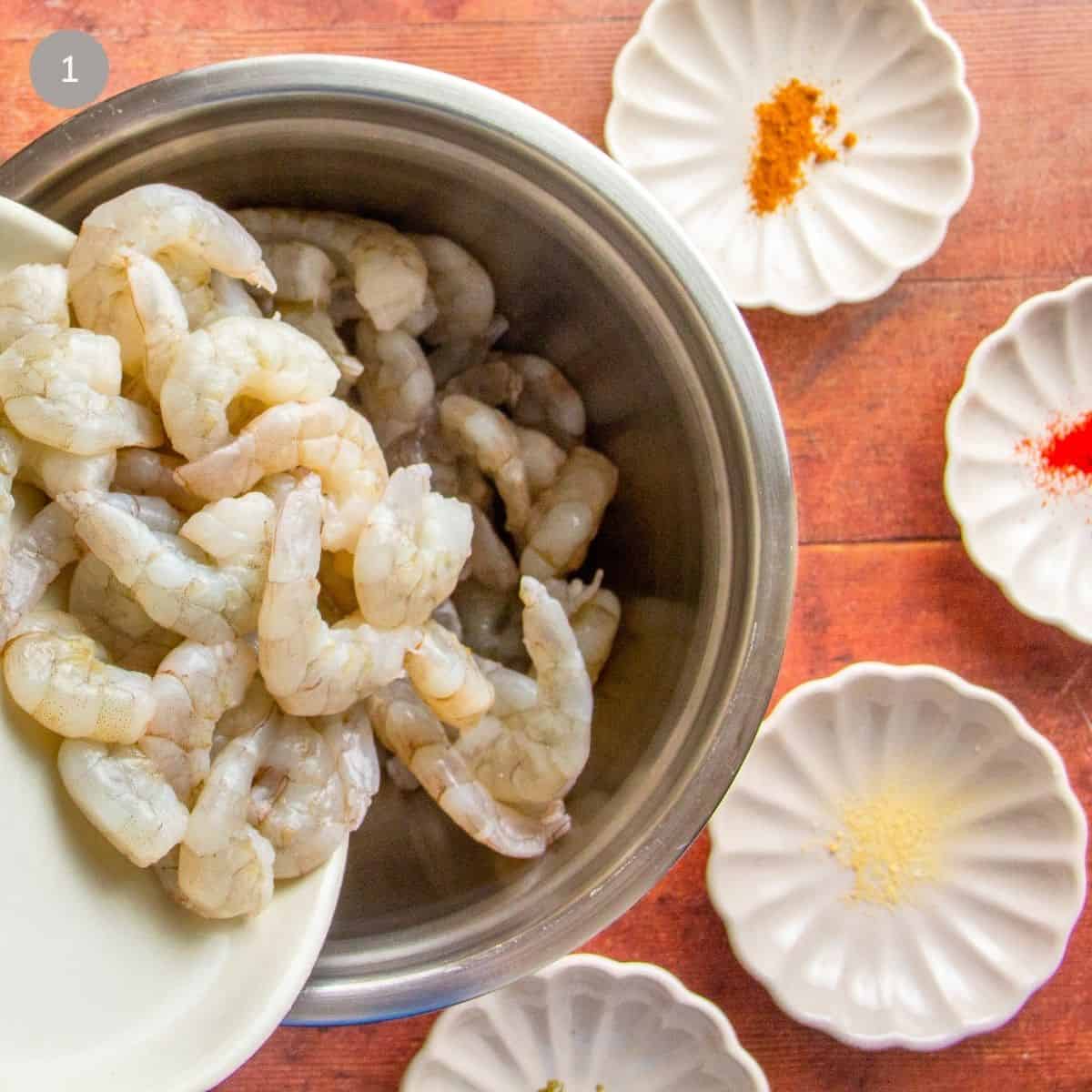 Raw shrimp in a metal bowl beside small dishes of cumin, chile powder, and garlic powder.