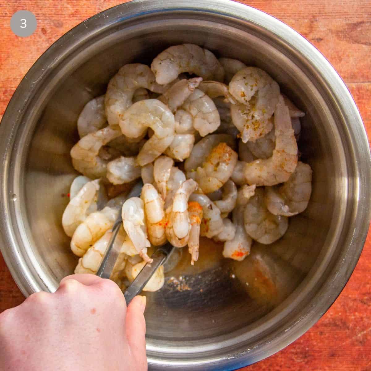 Tongs toss raw shrimp coated in spices and lemon juice in a metal bowl.
