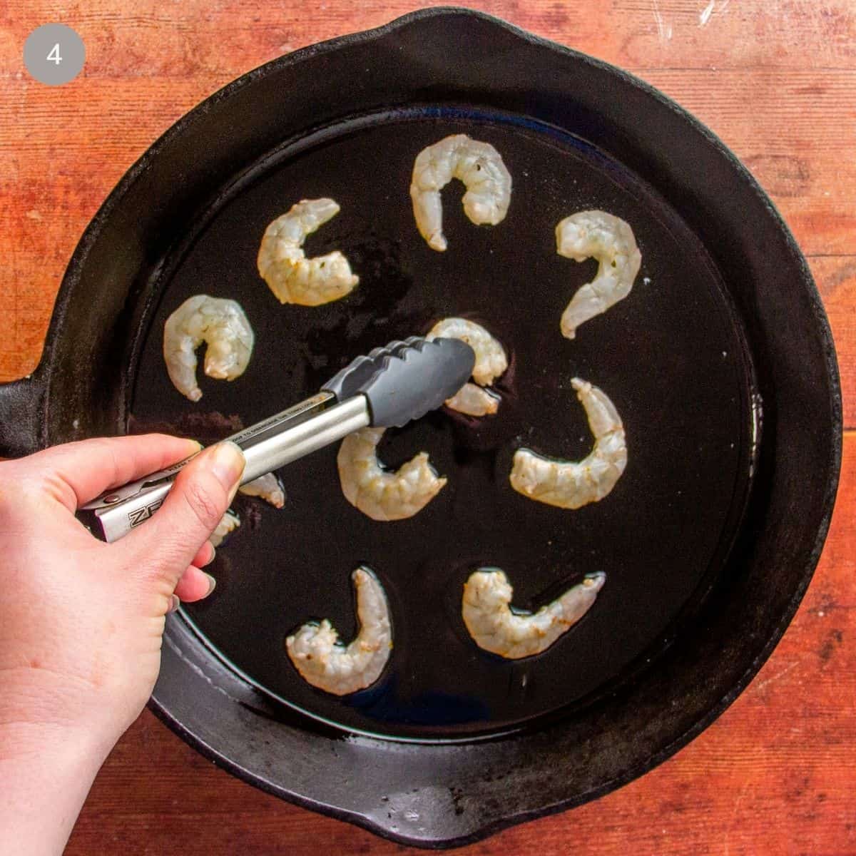 Raw shrimp arranged in a cast iron skillet, tongs turning one piece.