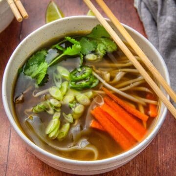 A bowl of broth holds carrots, bean sprouts, scallions, jalapeño, and cilantro with chopsticks resting across it.