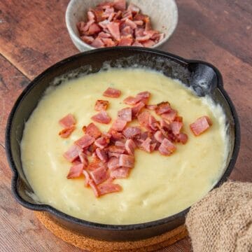 Melted cheese dip topped with bacon in a cast iron skillet, beside a bowl of bacon.