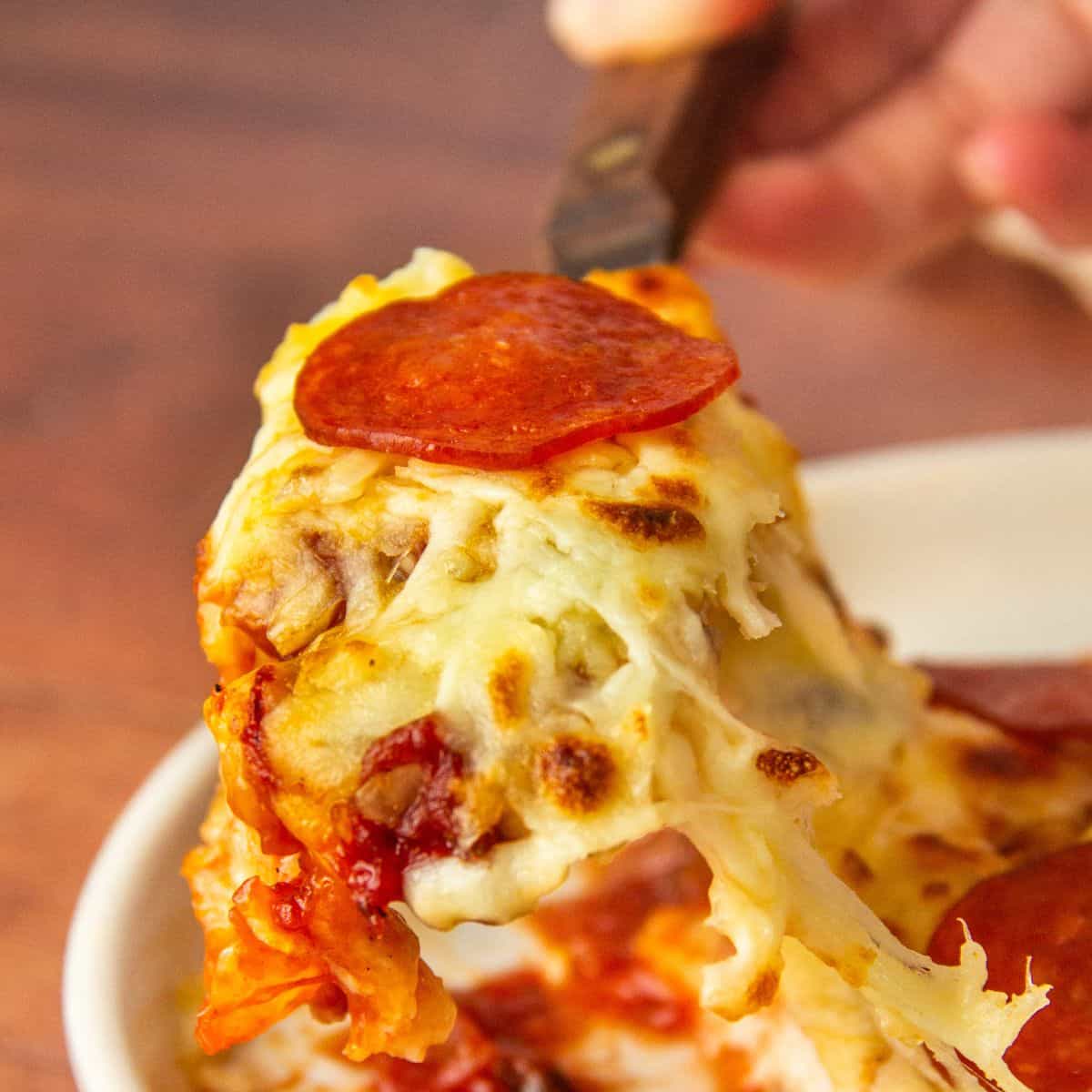 Fork lifts a cheesy, pepperoni-topped bite from a baked chicken breast in a white dish.