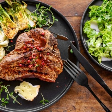Grilled T-bone steak topped with pomegranate seeds plated on a black plate with grilled fennel, a slice of brei cheese, and a side salad with fork and knife.
