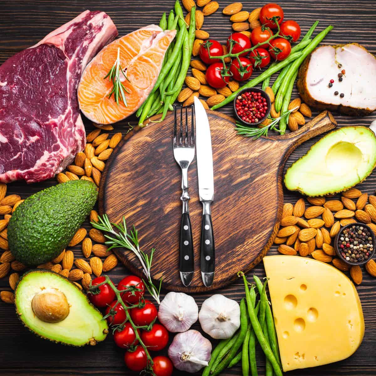 Knife and fork with black handle sitting on a wooden cutting board surrounded by cheese, garlic, avocado, steak, salmon, tomatoes, green beans, and spices.