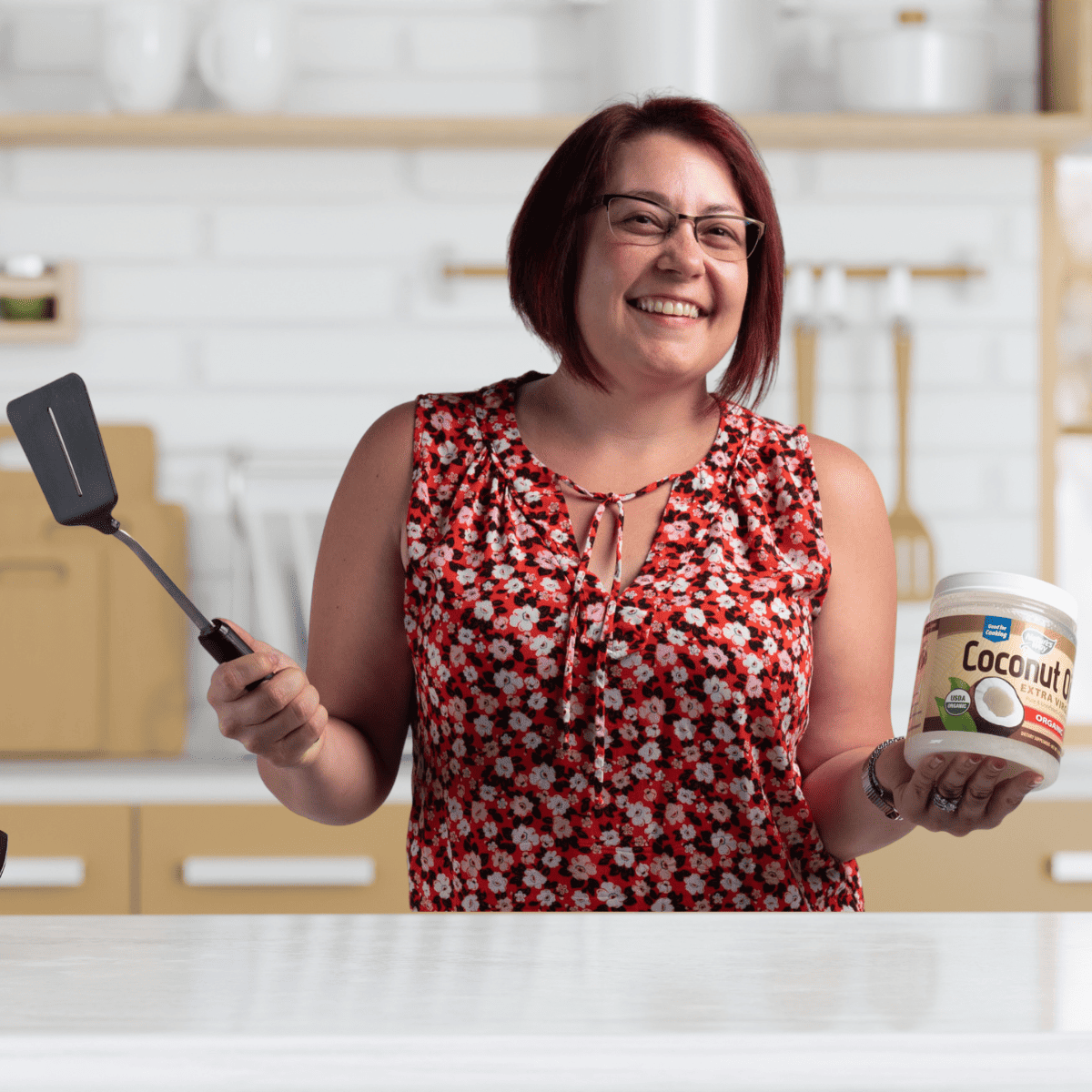 Heather Cooan in kitchen holding spatula and coconut oil wearing a red floral sleeveless shirt.