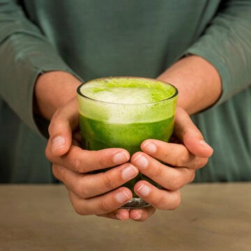 Woman's hands holding glass of green juice over a wooden countertop.