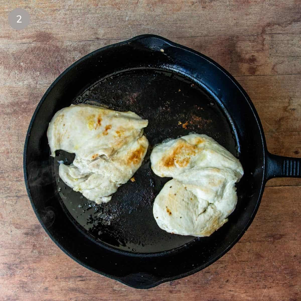 Chicken breasts browning in a cast iron skillet, golden on the edges and cooking in melted fat.