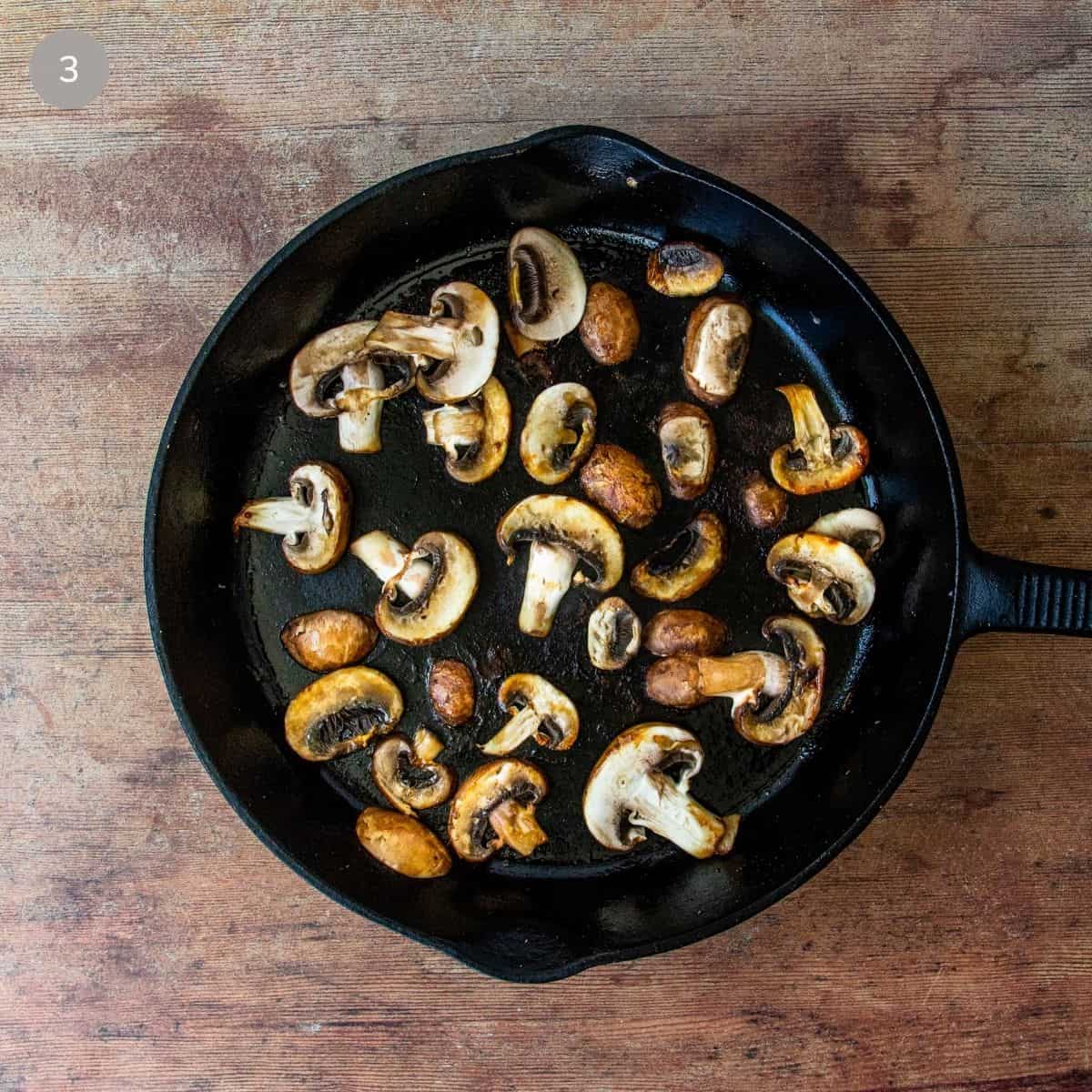 Sliced mushrooms browning in a cast iron skillet, golden and caramelized on the edges.